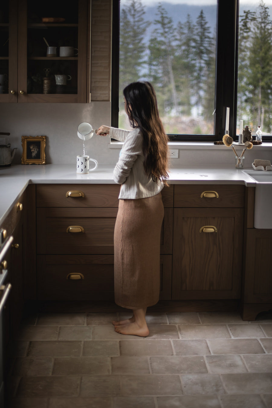 A kitchen floored in Italian Porcelain in Crema, 8x8 and 8x12 together, with walnut cabinets, brass pulls and a woman standing at the sink by the window