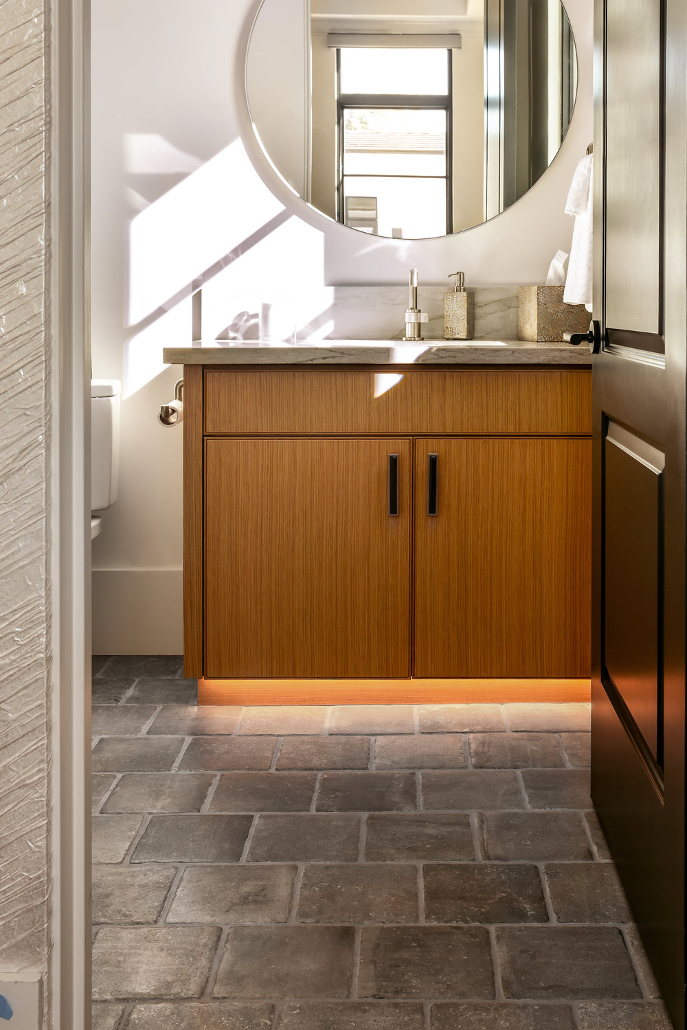 A bathroom floored in Italian Porcelain 8x8 in Grigio, laid in an irregular stone pattern, with a wood vanity, black pulls, a stone counter and a round mirror above it