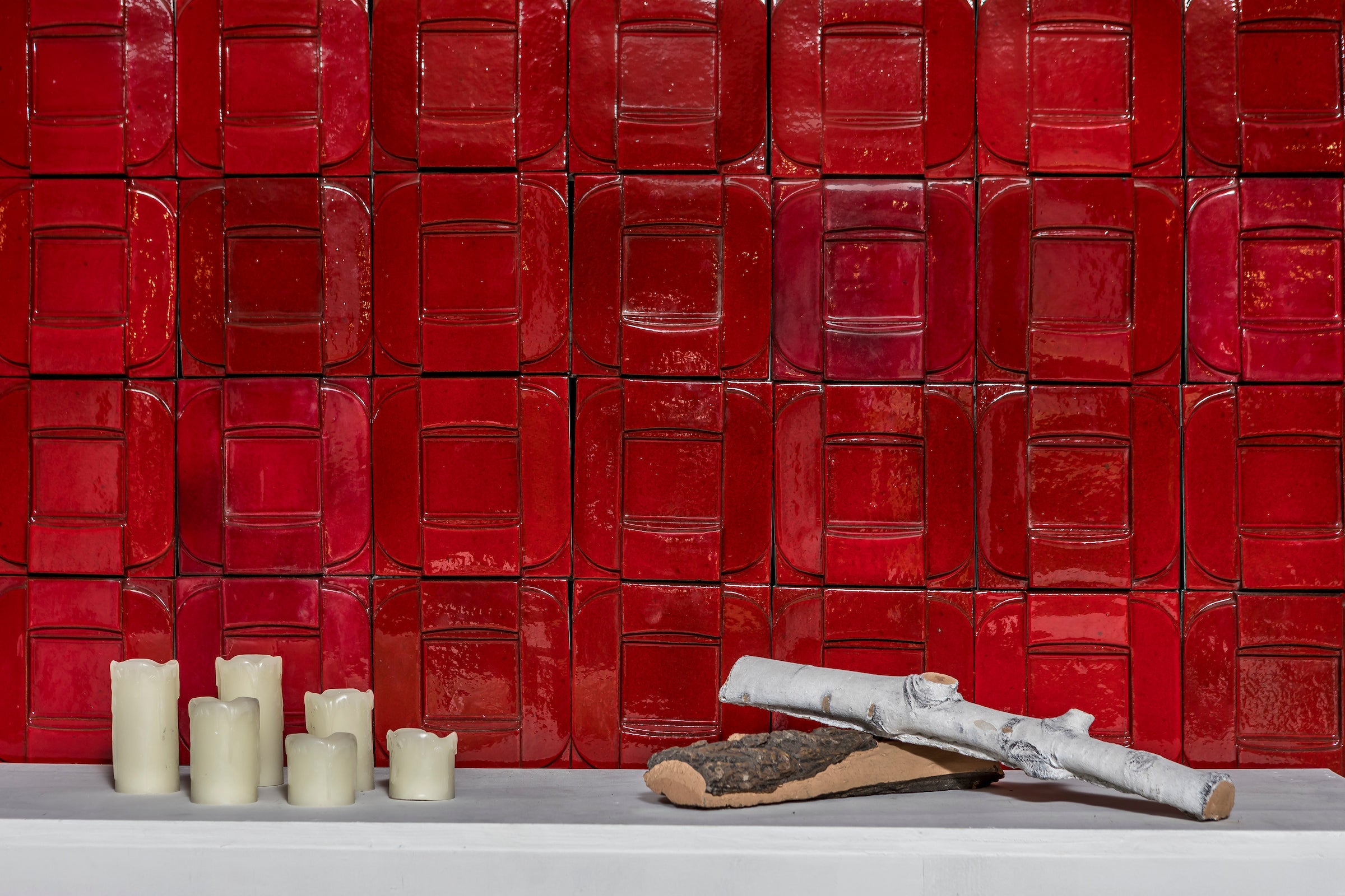 A wall of glossy red 3D relief tile above a white mantel, with candles and a birch log on the shelf