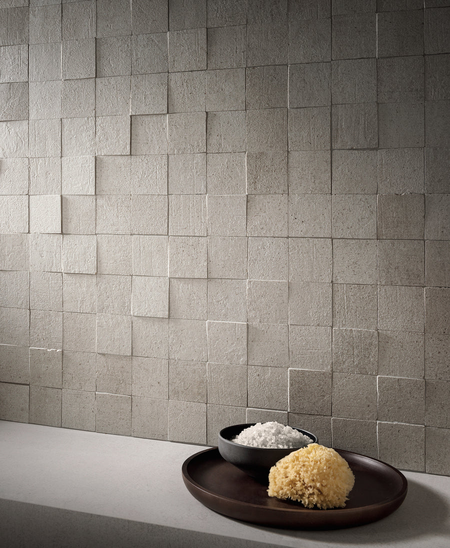 A wall of Terra Crea 3D porcelain mosaic in cool grey, lit from the side so every square stands off its neighbour, with a bowl of coarse salt and a natural sponge on the counter below