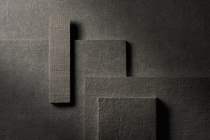 The same arrangement of Terra Crea pieces in Carbone, the darkest of the five, lit hard from the side against a dark ground