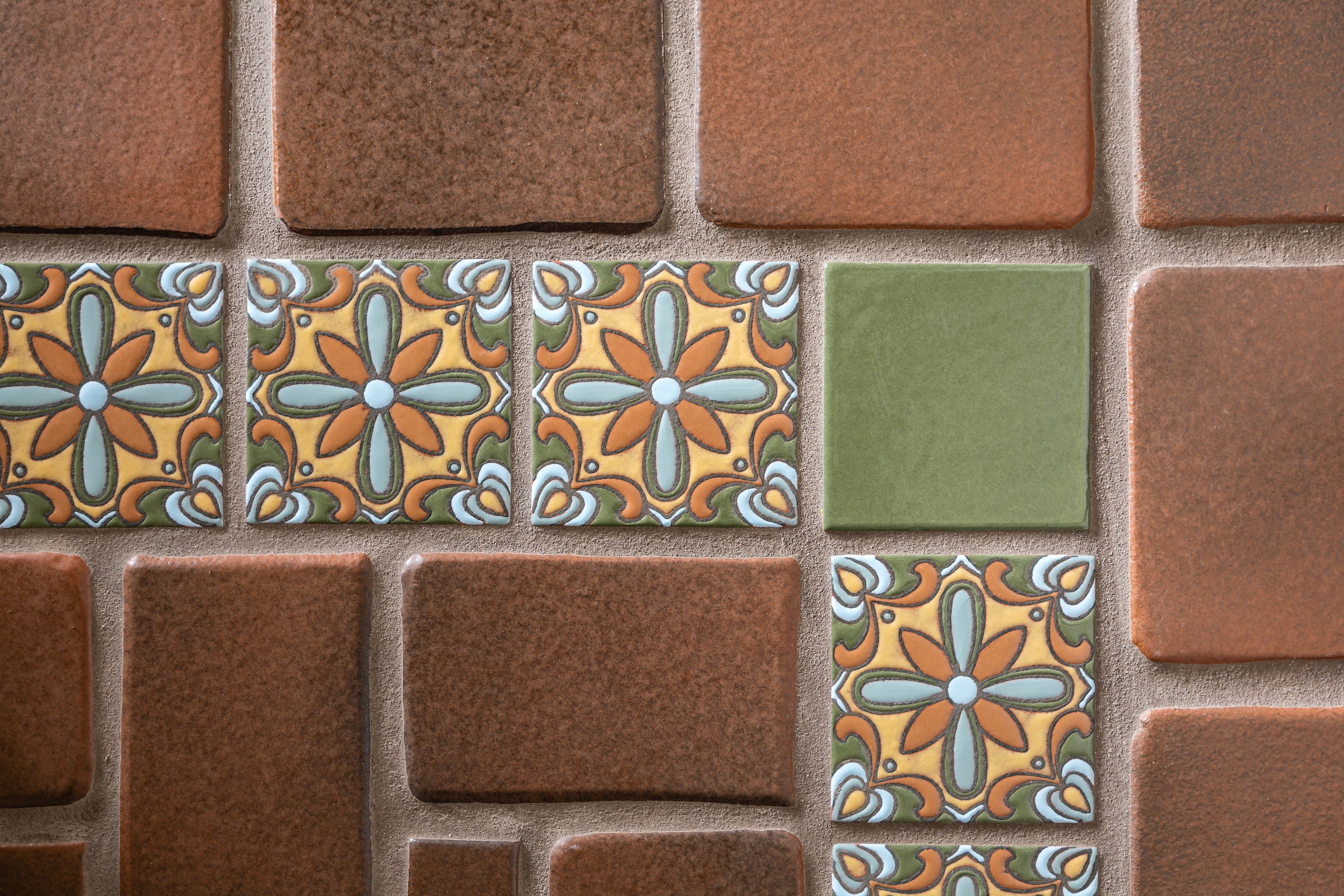 A wall of Oleson field tile in browns and one green, its pillowed edges catching the light, with a row of handpainted decos set into it