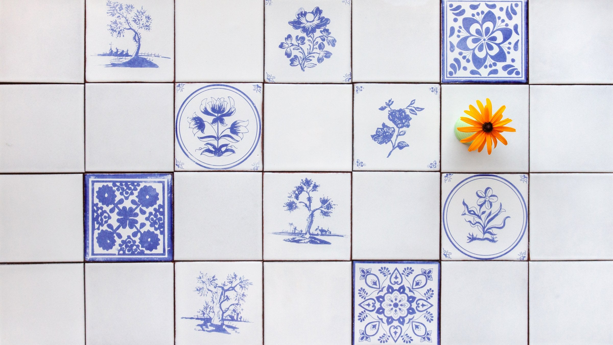 A wall of white Peninsula tile with blue drawings scattered through it, and a single orange flower in a small vase