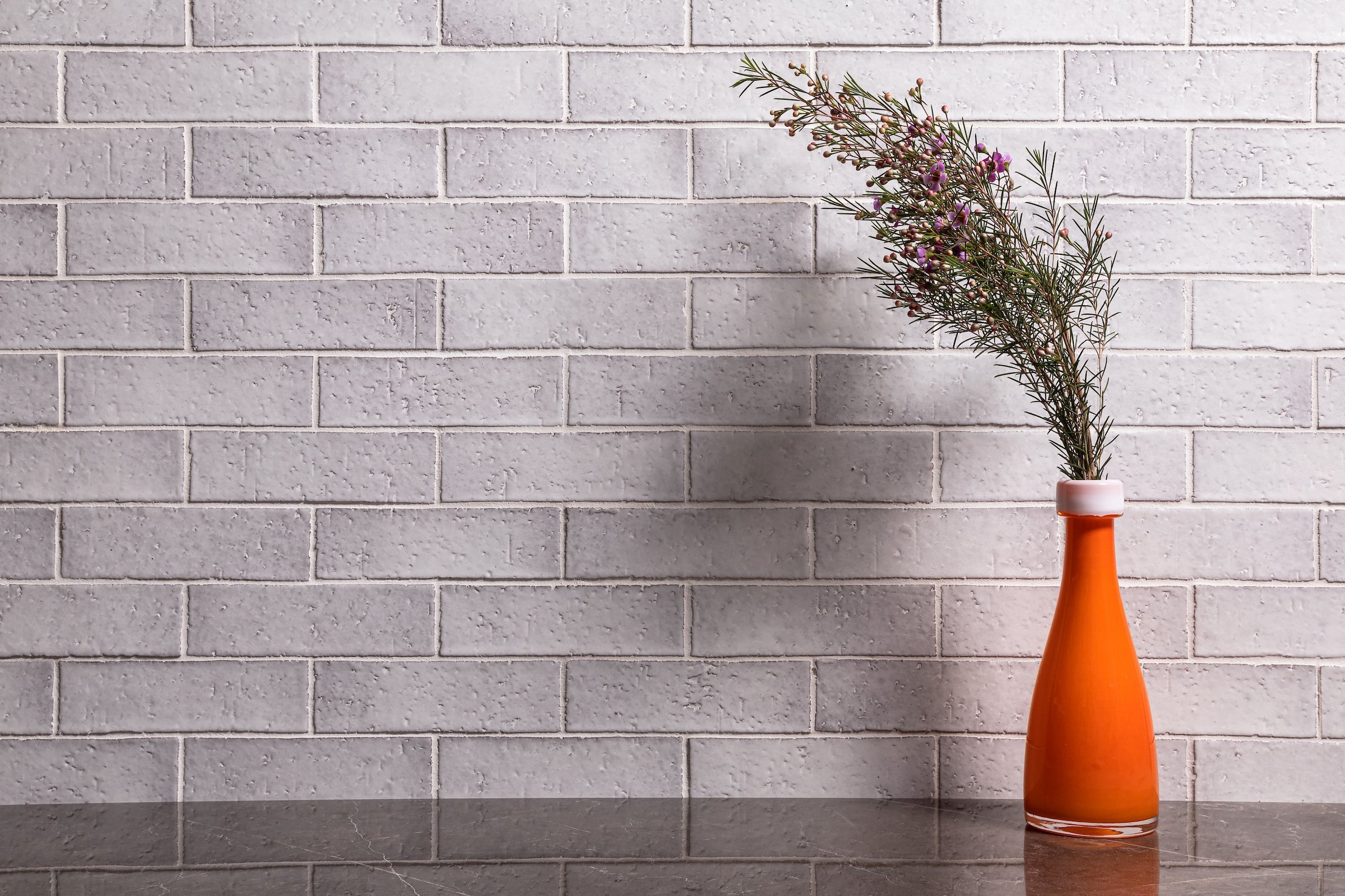 A wall of 2x8 glazed brick in Snowflake, laid in running bond, with a sprig of blossom in an orange bottle against it