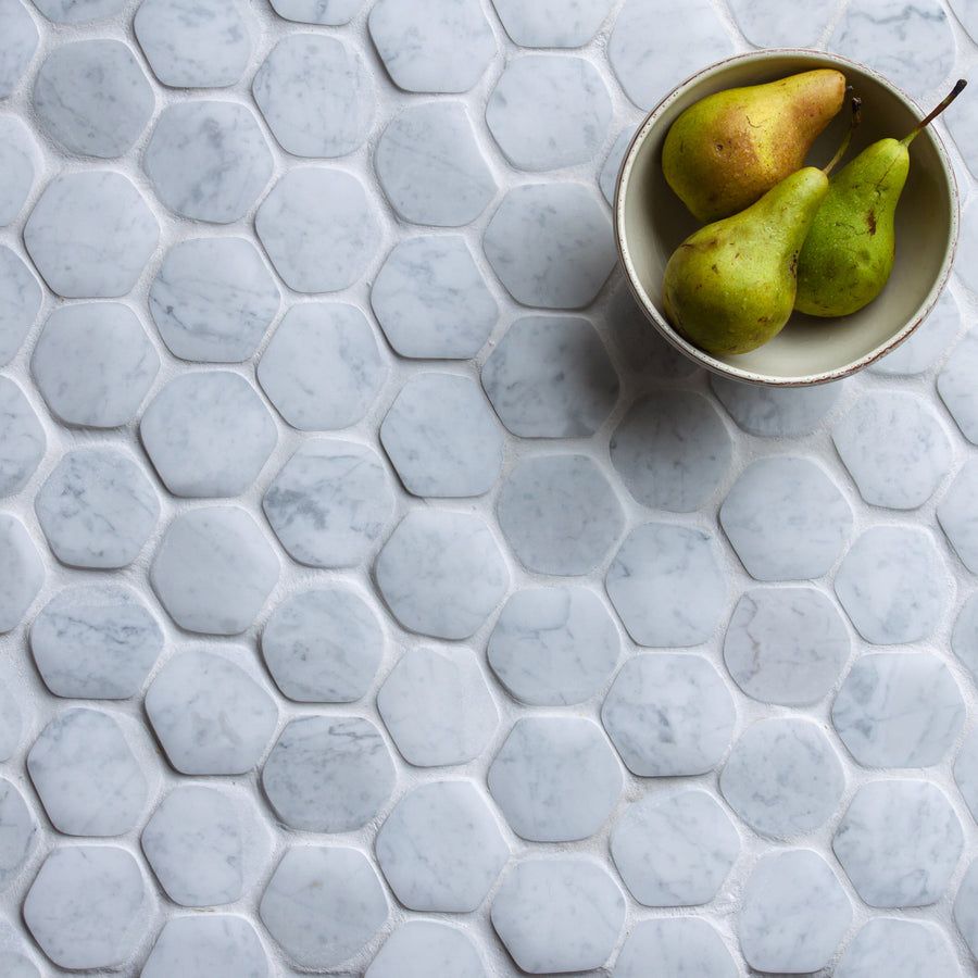 A hexagon marble mosaic floor seen from above in white and pale grey, with a shallow bowl of green pears resting on it