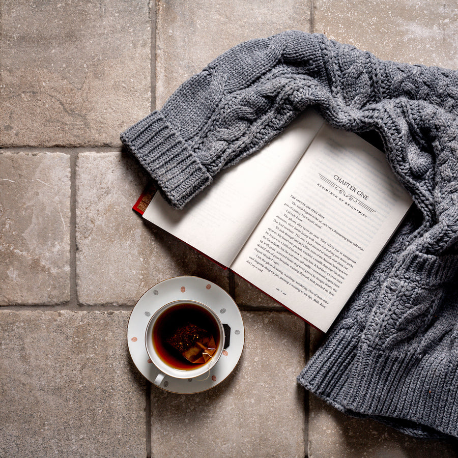 An Italian Porcelain floor in Crema, seen from above with an open book, a grey cable knit sweater and a cup of tea set down on it