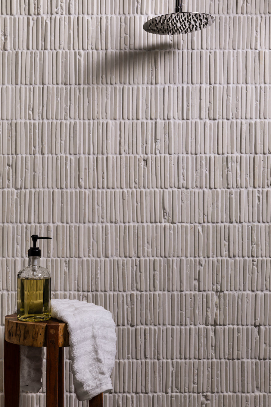 A shower wall tiled in narrow cream marble mosaic, a wooden stool with a soap bottle and a folded towel at its foot
